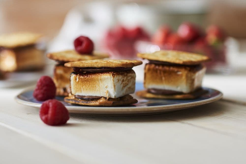 En tallrik med s'mores med rostade marshmallows och choklad mellan grahamskex, garnerade med hallon.