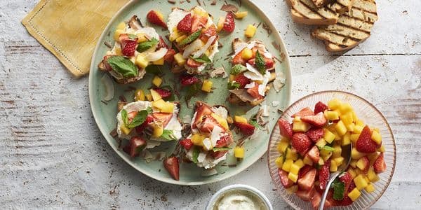 En tallrik med frukt-toppad bruschetta med jordgubbar, mango och mynta, bredvid en skål med blandad frukt, grillade brödskivor och en liten skål med grädde.