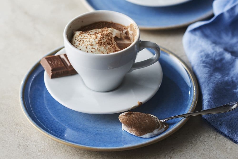 En kopp varm choklad toppad med vispgrädde och kakaopulver på en blå tallrik, tillsammans med en chokladbit och en sked.