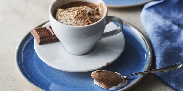 En kopp varm choklad toppad med vispgrädde och kakaopulver på en blå tallrik, tillsammans med en chokladbit och en sked.