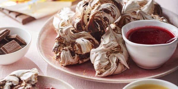 En tallrik med chokladvirvlade maränger, tillsammans med en skål röd sås och chokladbitar på en rosa yta.
