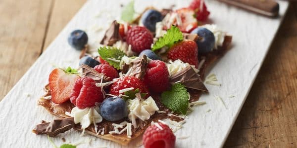 En dessert med hallon, blåbär, jordgubbar, chokladspån och myntablad på ett rektangulärt bakverk, serverat på en vit tallrik.