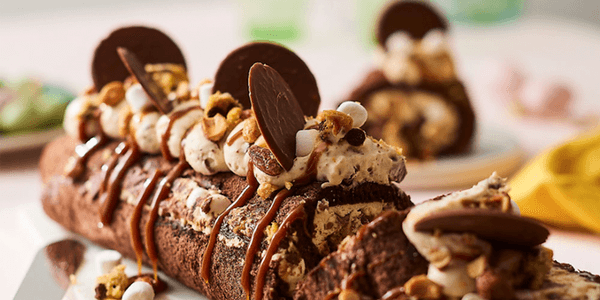 Chokladrulltårta toppad med vispgrädde, kolasås, chokladdiskar och små marshmallows på ett vitt serveringsfat.