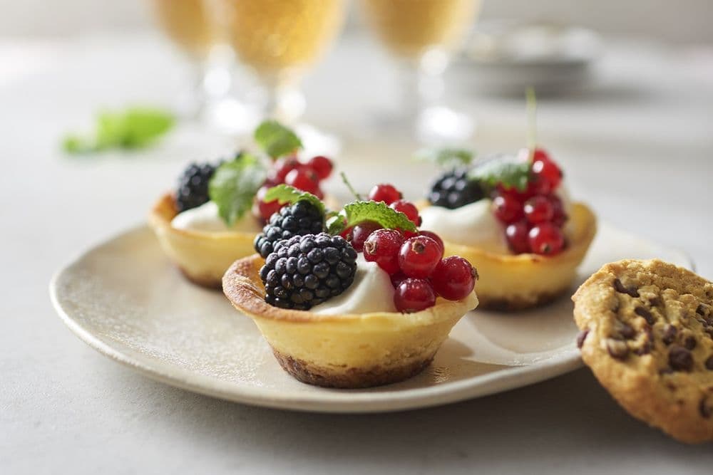Mini-cheesecakes toppade med björnbär, röda vinbär och myntablad på en tallrik, med en chocolate chip-cookie och drycker i bakgrunden.