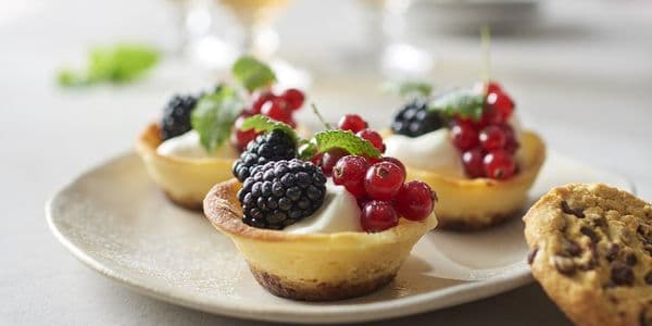 Mini-cheesecakes toppade med björnbär, röda vinbär och myntablad på en tallrik, med en chocolate chip-cookie och drycker i bakgrunden.