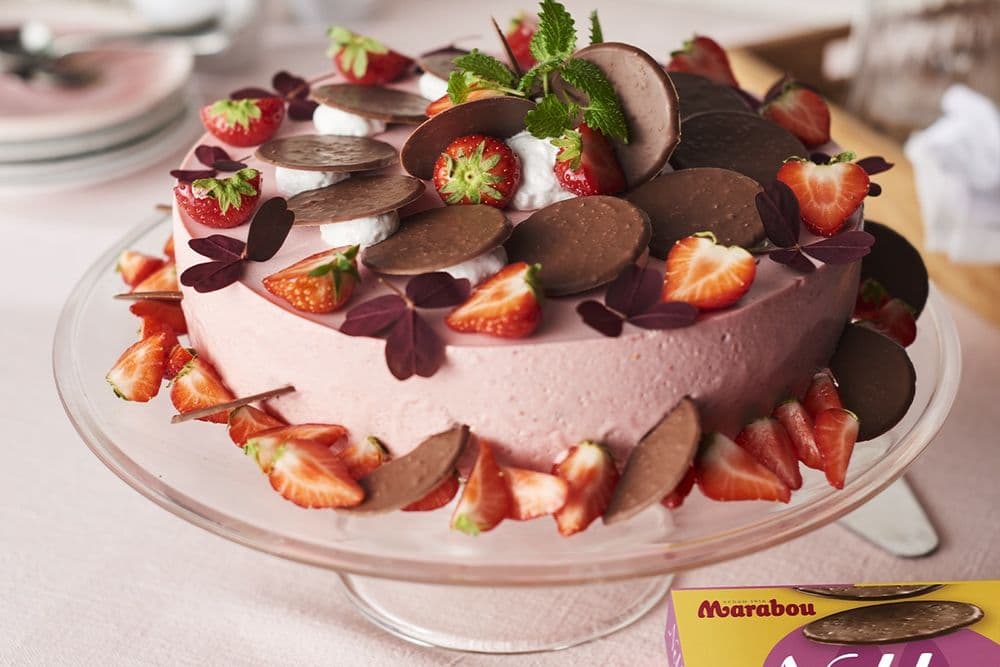 En rosa tårta toppad med chokladdiskar, jordgubbar och gröna blad på ett glasfat, med en ask Marabou-choklad intill.