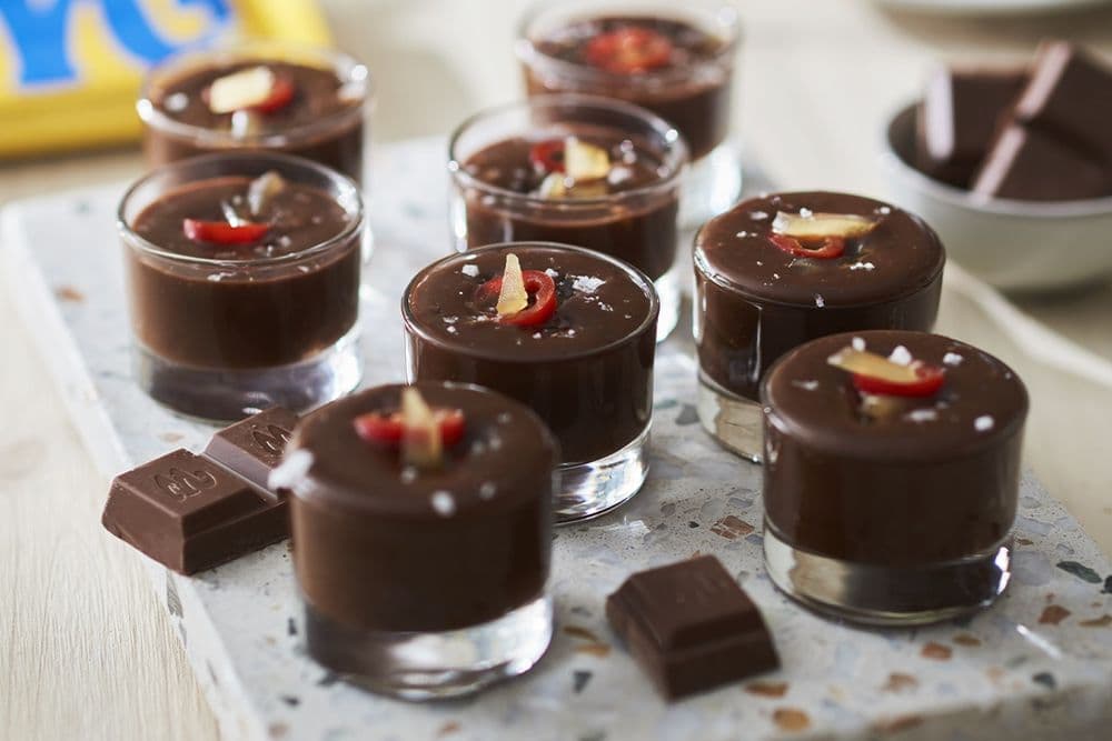Små glas med chokladmousse toppade med skivade chilifrukter och havssalt, arrangerade på en stenskiva med chokladbitar intill.