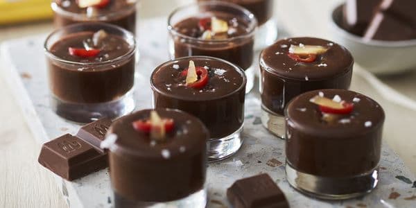 Små glas med chokladmousse toppade med skivade chilifrukter och havssalt, arrangerade på en stenskiva med chokladbitar intill.